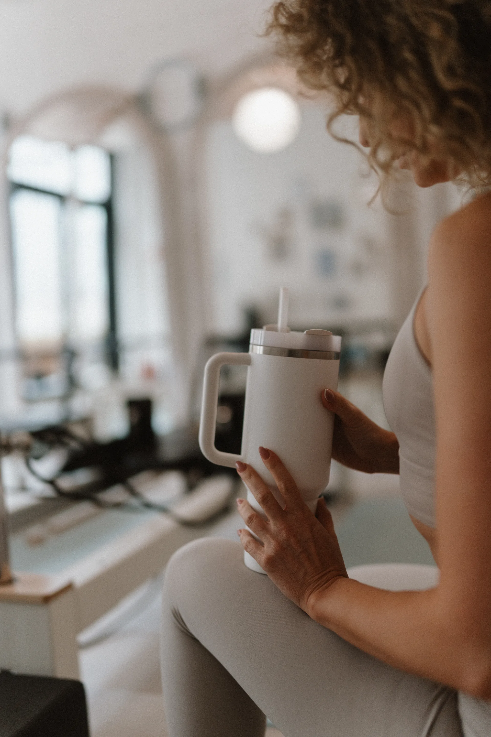 Pilates’nfinity reformer stúdióban ülő vendég világos sportruhában, kezében fehér kulaccsal, elmosódott stúdióháttérrel.