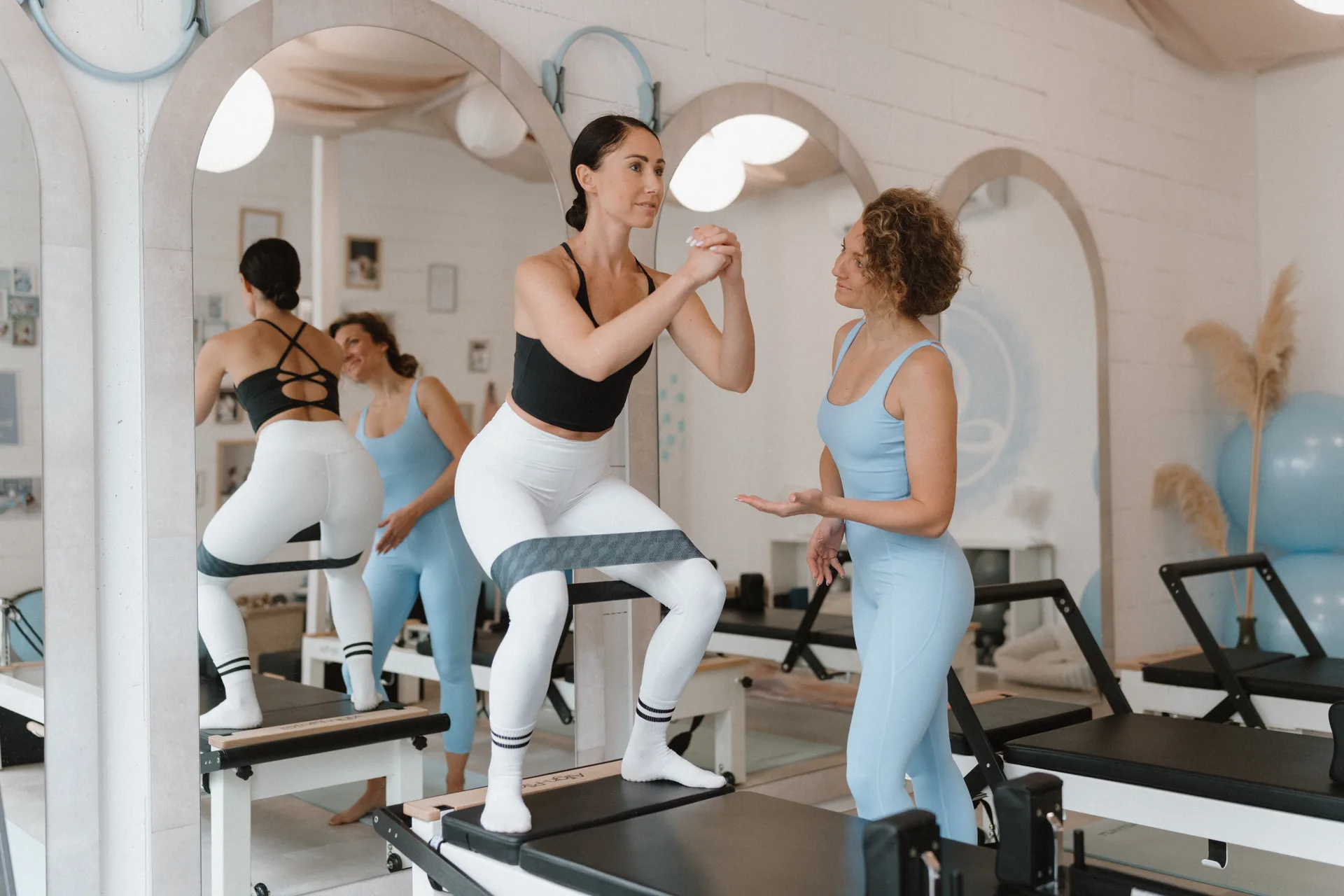 Reformer pilates gyakorlat térdvalgus kontrollra – vendég gumiszalaggal guggol a Pilates’nfinity budaparti stúdiójában, oktatóval a háttérben.