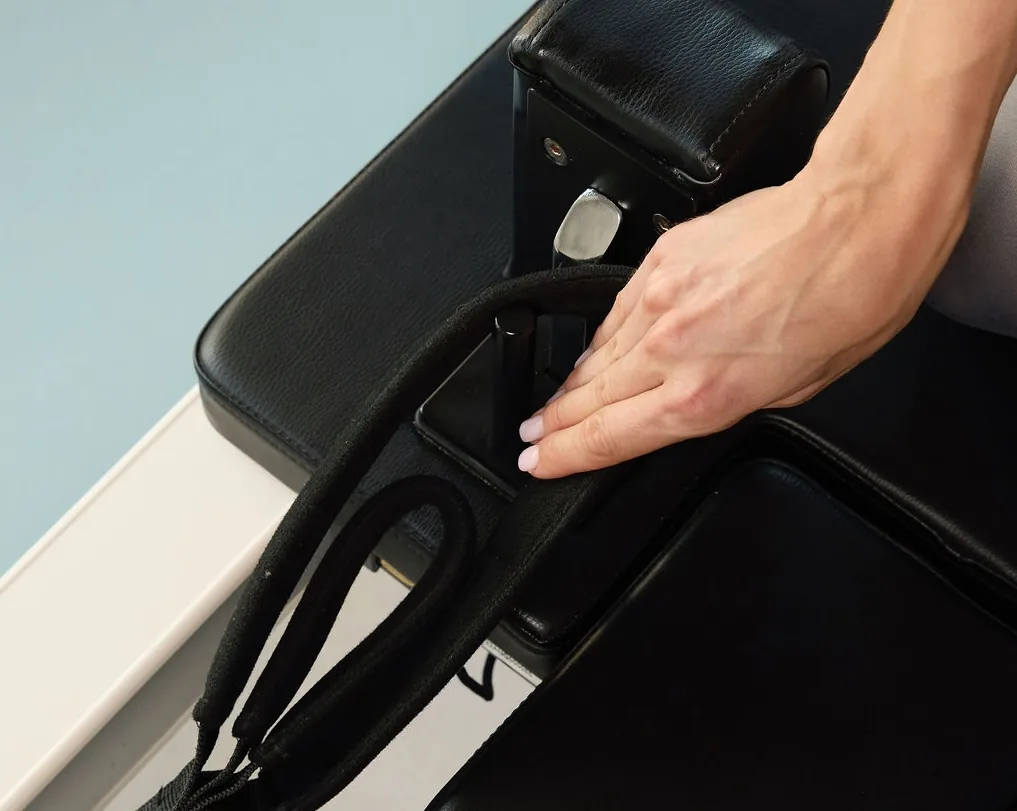 Közelkép egy kézről, amint a reformer gép hevederét állítja a Pilates’nfinity stúdióban.