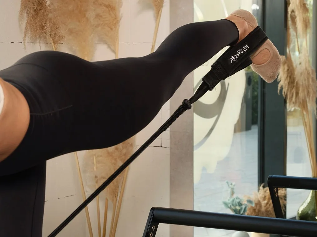 Reformer strapben dolgozó láb és talp közelről, lábboltozat-erősítő gyakorlat a talp–térd–csípő tengely stabilizálására a Pilates’nfinity budaparti stúdiójában.