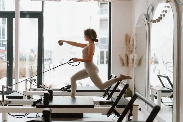 Reformer pilates gyakorlat: álló kitörés és kontrollált karfeszítés egy prémium, napfényes stúdiókörnyezetben.